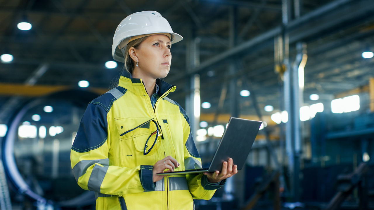 Female Industrial Engineer in the Hard Hat Uses Laptop Computer while Standing in the Heavy Industry Manufacturing Factory. In the Background Various Metalwork Project Parts Lying 