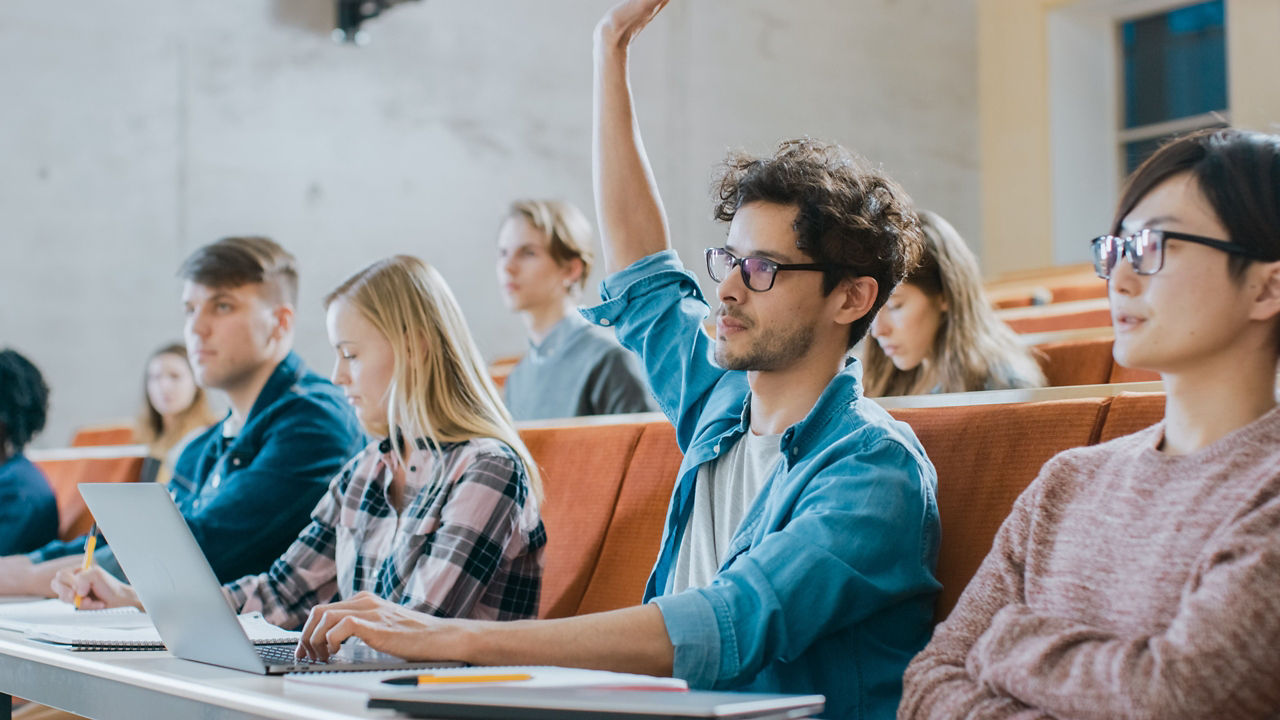 Handsome Hispanic Student Uses Laptop while Listening to a Lecture at the University, He Raises Hand and Asks Lecturer a Question. Multi Ethnic Group of Modern Bright Students.