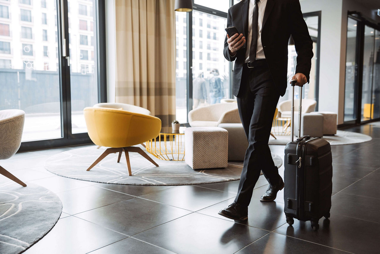 Cropped photo of successful businessman wearing suit holding smartphone and walking with suitcase in hotel lobby