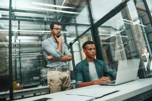 Busy day. Two young and handsome men in casual wear looking at screen of laptop and thinking about new project while working in modern office
