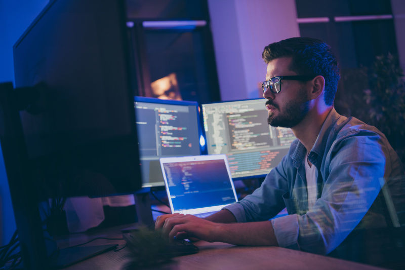 Man working in a dark office monitoring multiple monitor screens