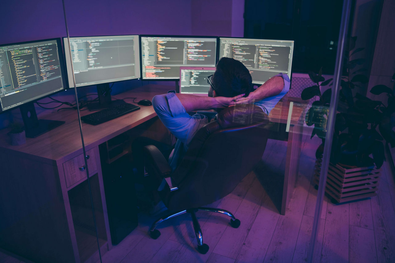 Rear back behind view of nice serious skilled genius brunet guy sitting in chair monitoring creating data database information hardware solution consultant in dark violet room workplace station