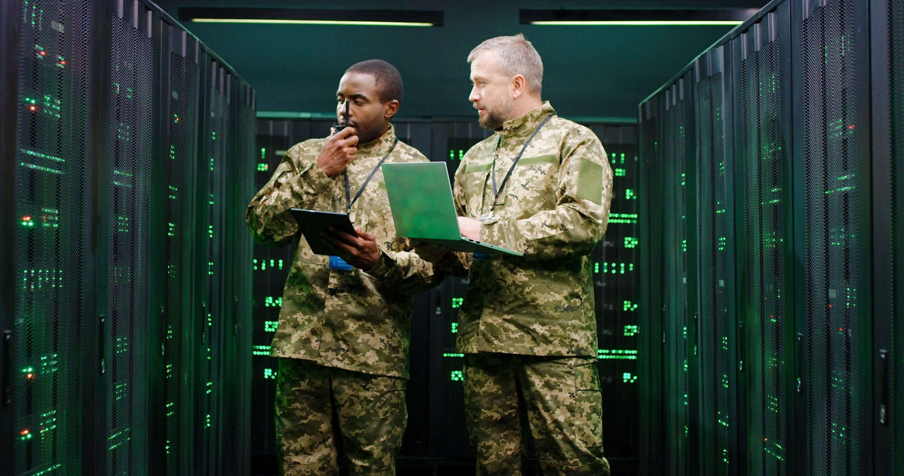 Multiethnic male army officers with tablet device and laptop computer checking secret data in server room and passing via walkie-talkie. African American and Caucasian men talking at digital facility.