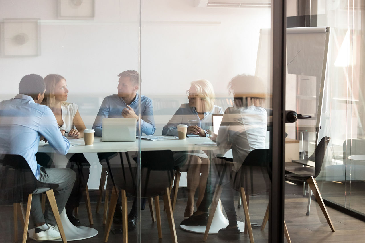 Team of network professionals seated around a conference table in a glass-walled meeting room engaged in discussion with laptops