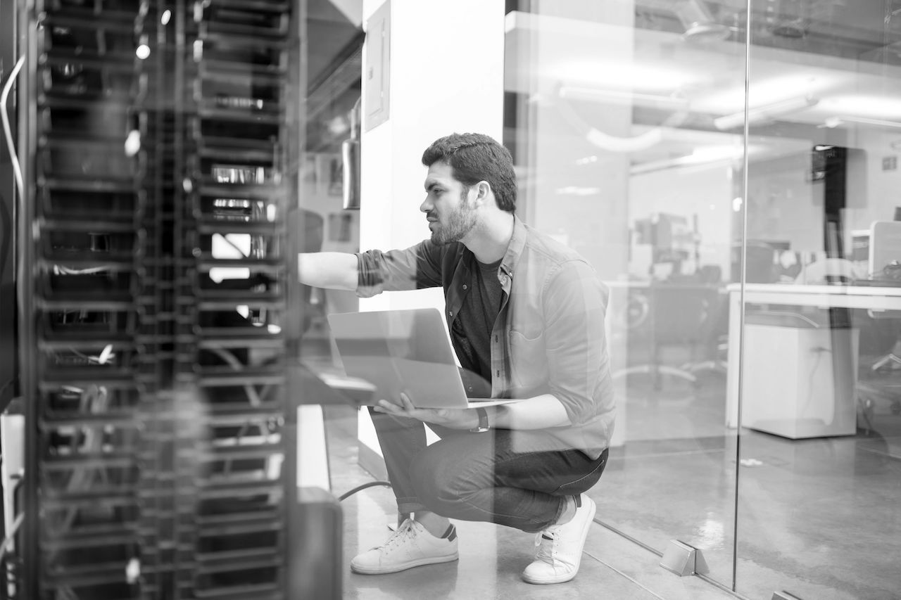 Black and white image of network administrator with notebook computer sitting in data center room and working with networking device on rack cabinet