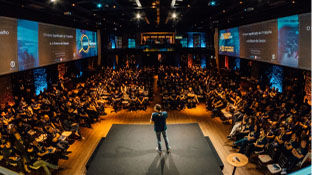A keynote speaker giving a presentation at a conference. There are dozens of people sitting around them. The presentation is displayed via projector on all the walls in the room.