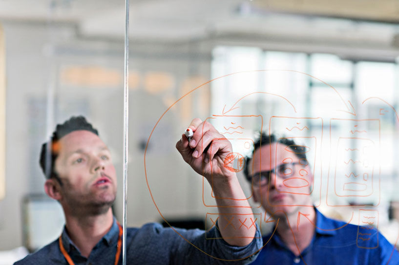 オレンジ色のマーカーでガラス壁に書き込んでいる2人の開発者。