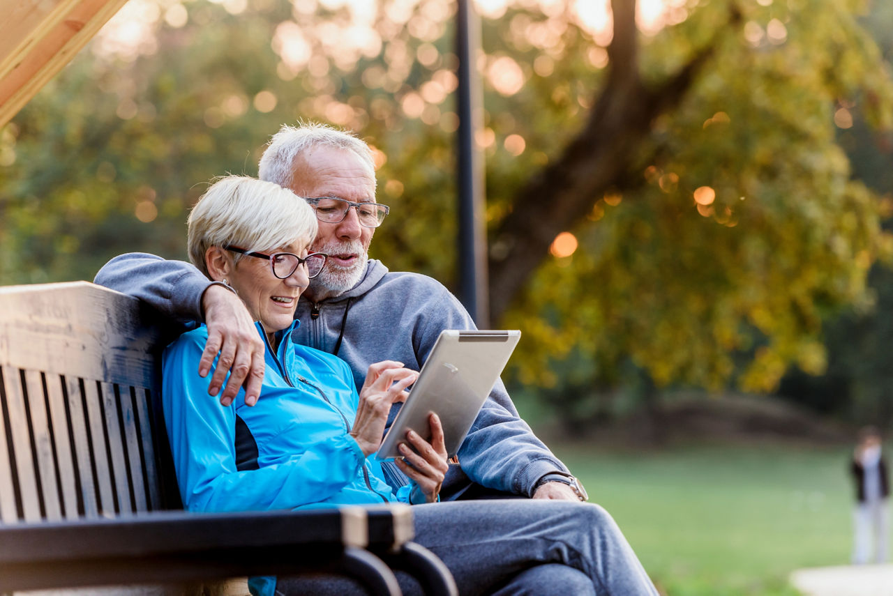 Smiling senior active couple sitting on the bench looking at tablet computer. Using modern technology by elderly.