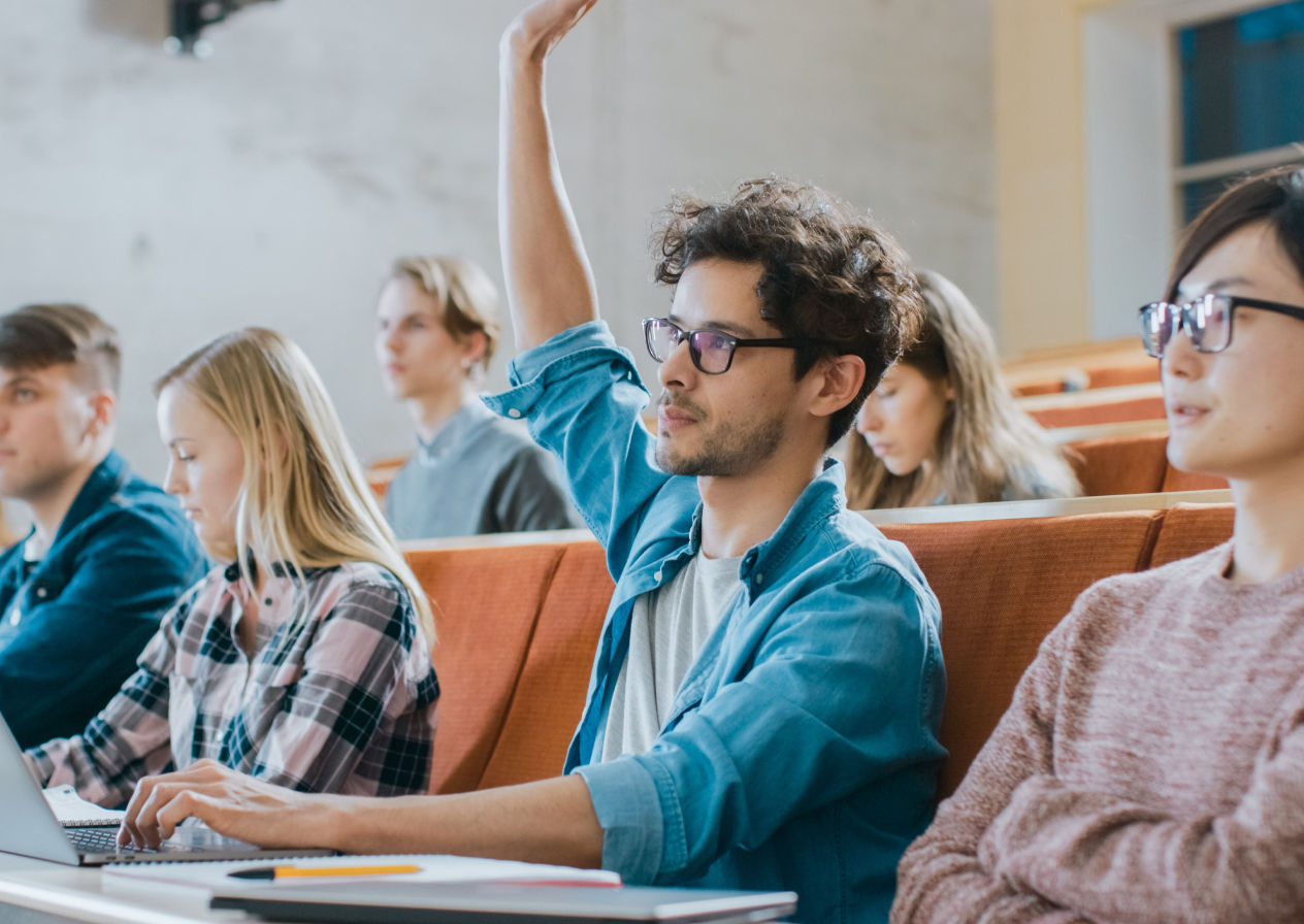 Handsome Hispanic Student Uses Laptop while Listening to a Lecture at the University, He Raises Hand and Asks Lecturer a Question. Multi Ethnic Group of Modern Bright Students.